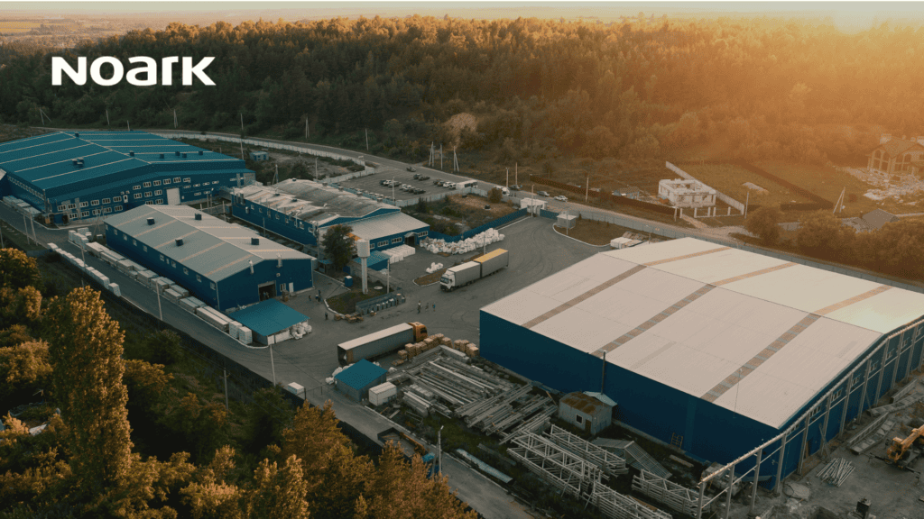 From above, the sprawling industrial complex unfolds—a network of blue-roofed buildings nestled among trees and intersected by a winding road. As the sun sets, the facility's molded case switches ensure smooth operations continue seamlessly into twilight.