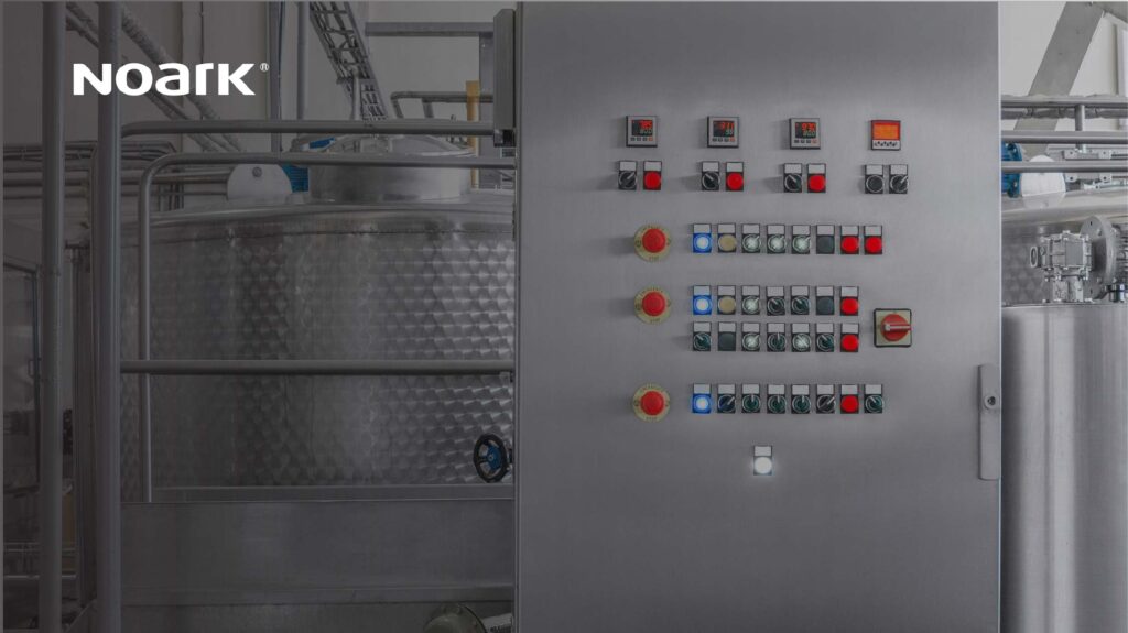 Industrial control panel with red and black buttons, switches, and indicators, including a motor starter for efficient operation. A large metal tank is visible in the background.