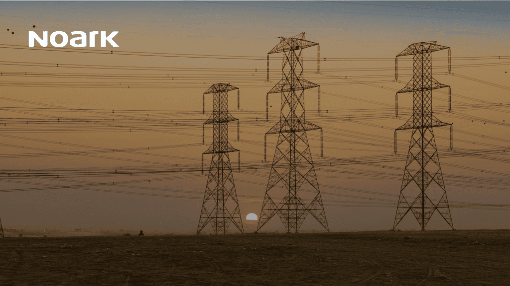 Three electricity pylons stand in a barren landscape at sunset, with numerous power lines and control circuit devices subtly integrating across the sky.