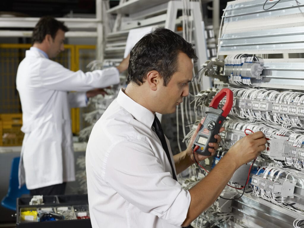 Two technicians work on electrical panels; one uses a multimeter, while the other inspects wiring. They are in a lab environment, wearing white shirts.