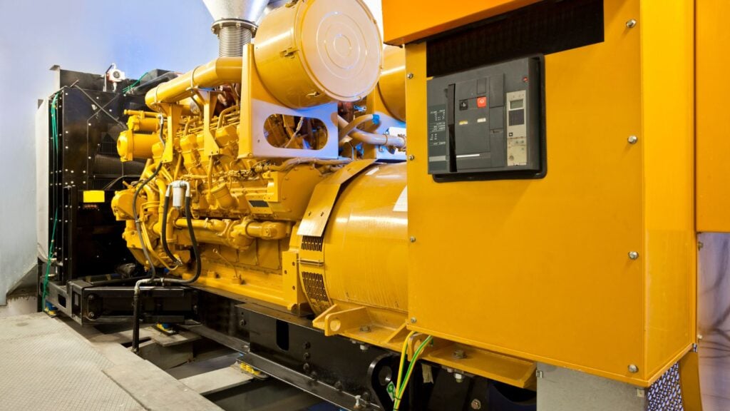 A large, industrial yellow generator with a robust engine is installed indoors, featuring a control panel and various cables.