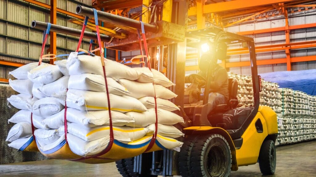 A forklift efficiently handles material by transporting a load of white sacks with a red lifting strap inside a warehouse supported by orange and steel beams.