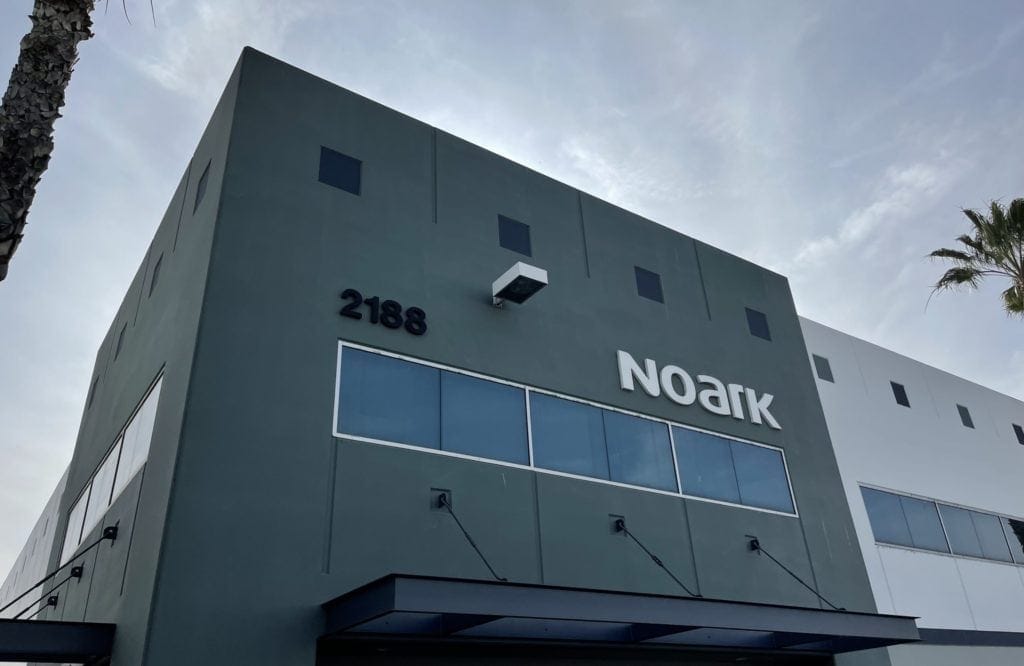 Two-story building with "Noark" logo and number 2188 on the facade. The structure has a modern design with large windows and a teal exterior. Sky and palm tree partially visible.
