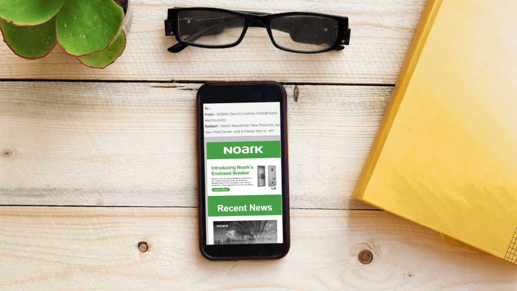 A smartphone electrifies the scene as it displays an email from NOARK on a wooden table, surrounded by glasses, a plant, and a yellow folder—a curious mailbox of modern communication.