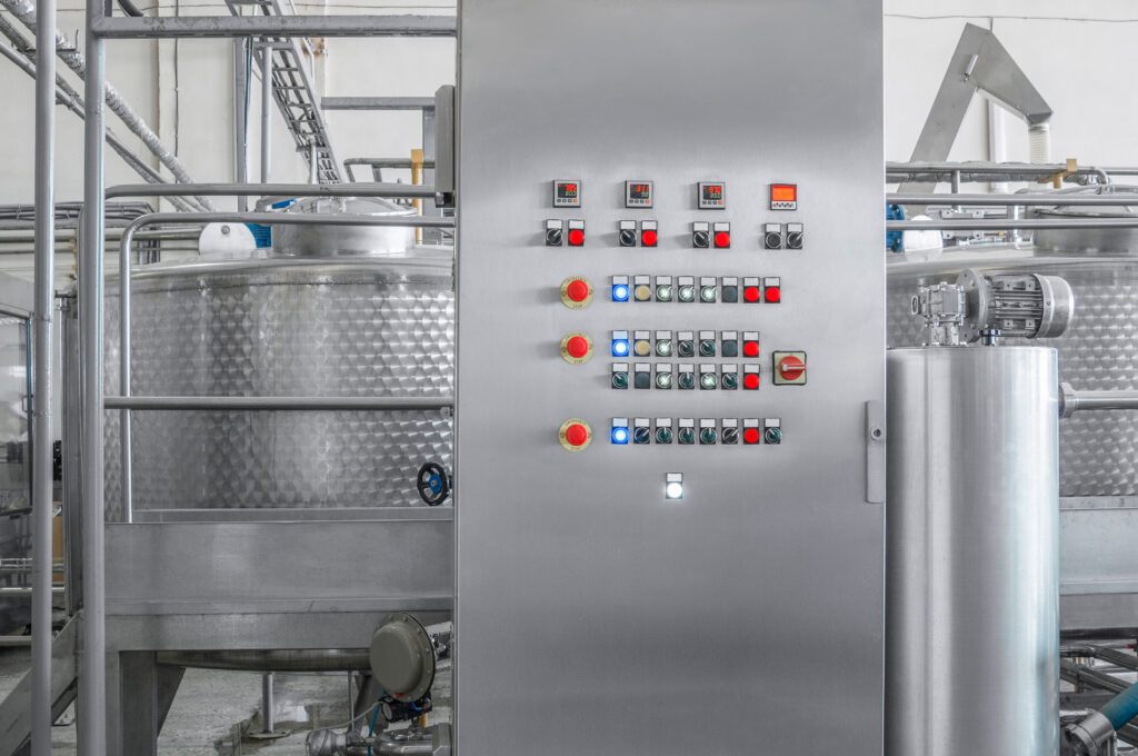 Industrial control panel with various buttons and switches, expertly crafted by a skilled panel builder. It's set against a backdrop of large stainless steel tanks and high-end equipment, meeting UL508A standards for safety and reliability.
