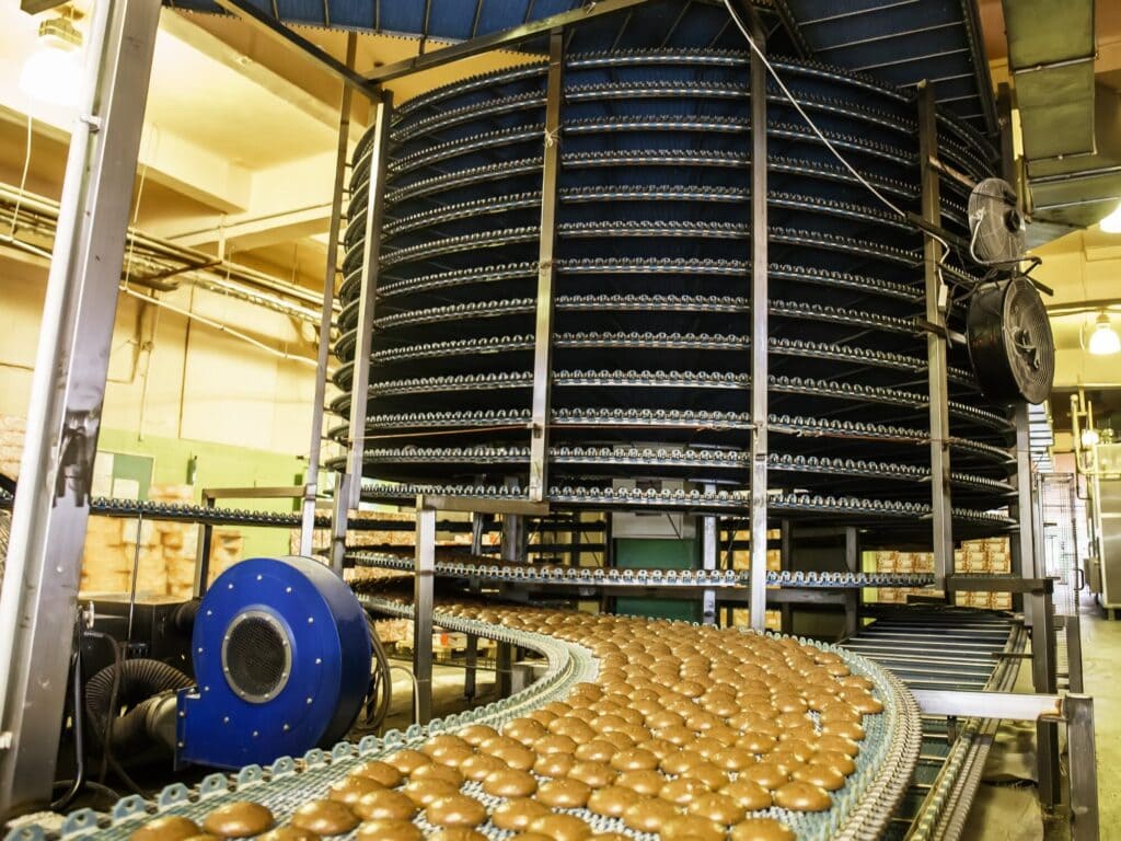 Rows of bread buns on a conveyor belt in a factory setting, with a large spiral cooling rack in the background.
