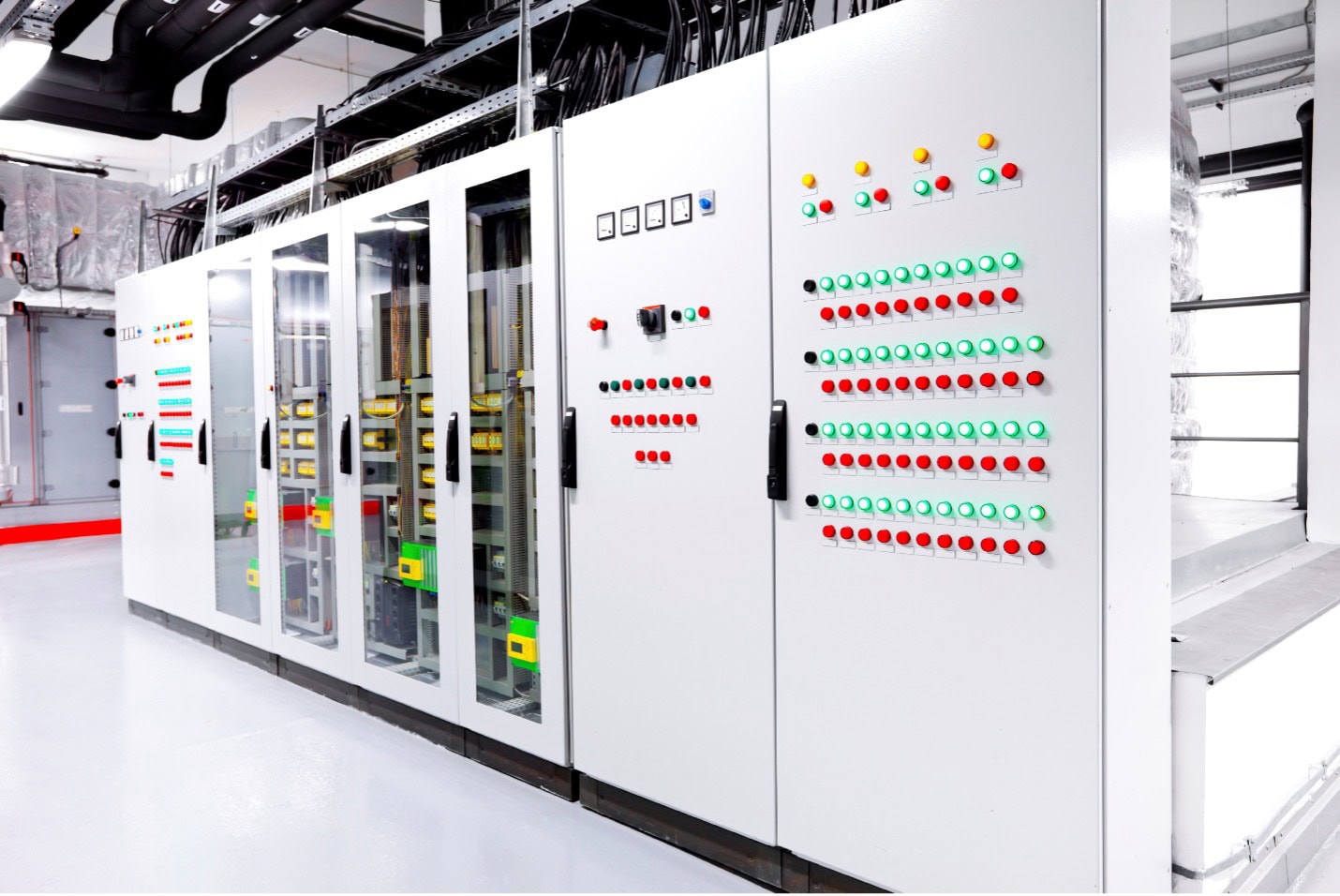 A row of white electrical control panels with various colored buttons, switches, and small displays in a brightly lit industrial setting.