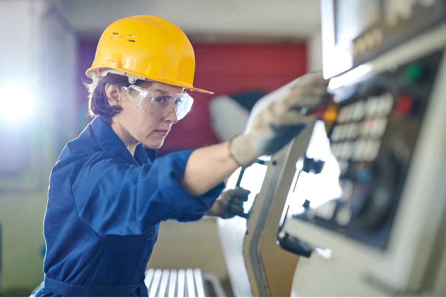 Person in a yellow hard hat and safety goggles operates a control panel in an industrial setting. They're wearing blue work attire and gloves.