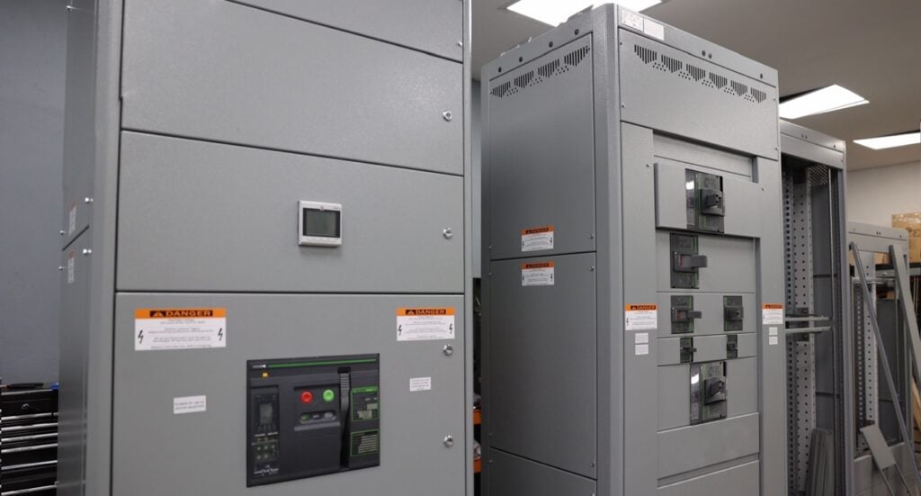Two large grey electrical cabinets, resembling industrial switchboards, feature control panels and caution labels in a well-lit room.