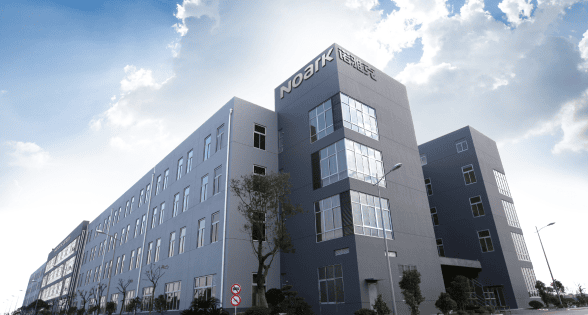 Large, modern industrial building labeled "NOBIK" with multiple windows and trees in front, under a partly cloudy sky.